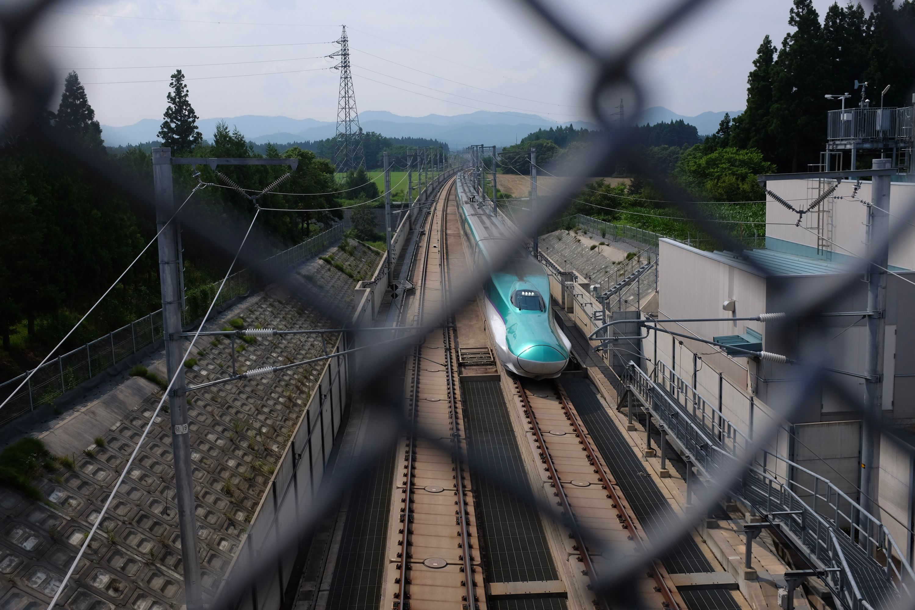 A light blue superexpress train, the Hokkaido Shinkansen, as seen from behind the fence of an overpass.