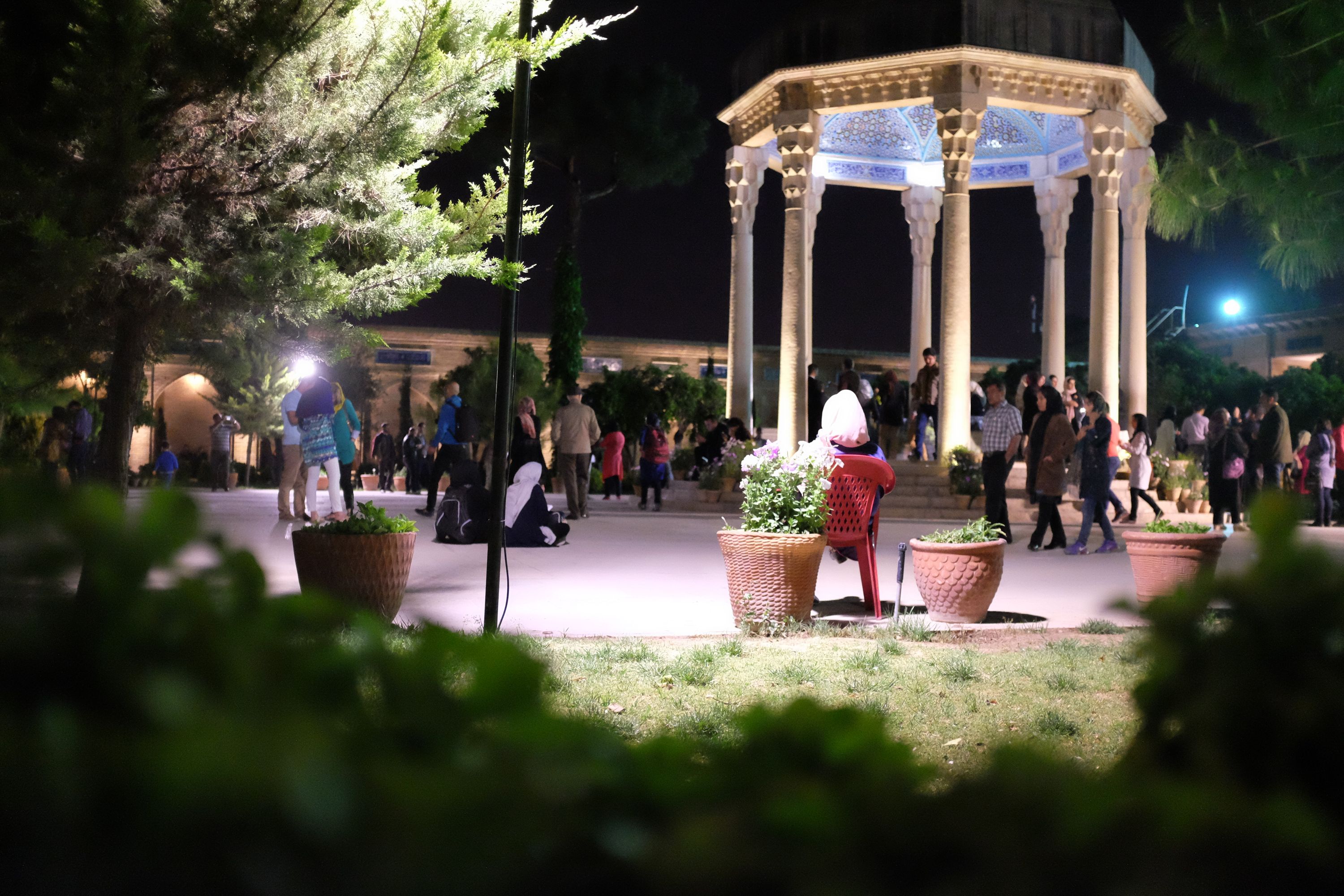 At night, people walk around an octogonal pavilion, framed by trees.