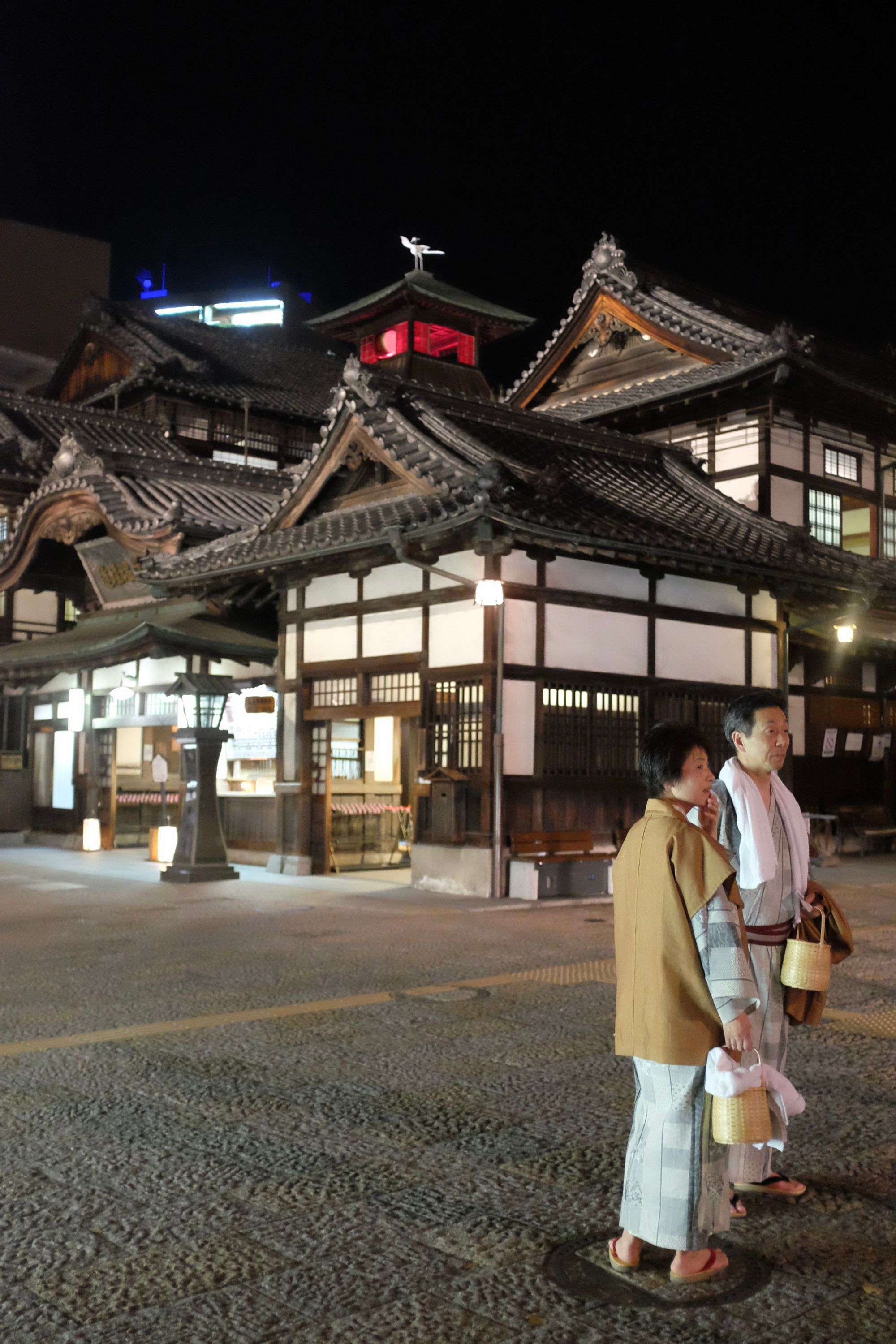 A Japanese couple, dressed in yukata and carrying bathing utensils, stands in front of the famous main building of Dōgo Onsen in the night.