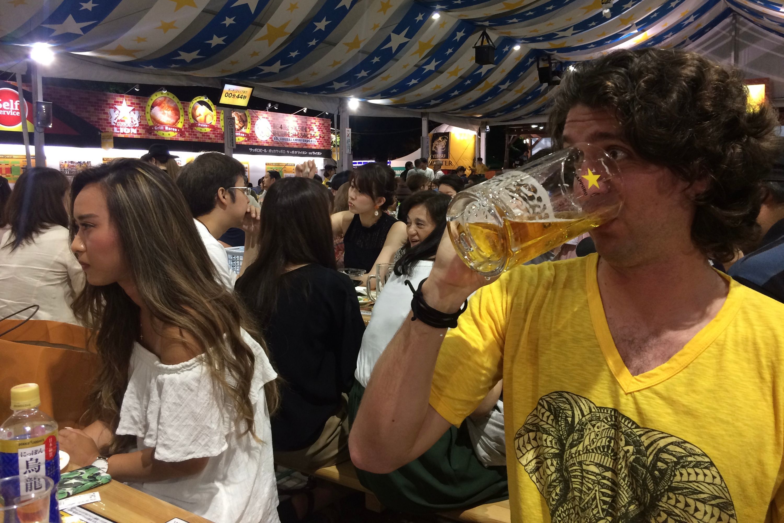Gabor drinks beer from a very large mug, sitting next to a Japanese woman in a white shirt.