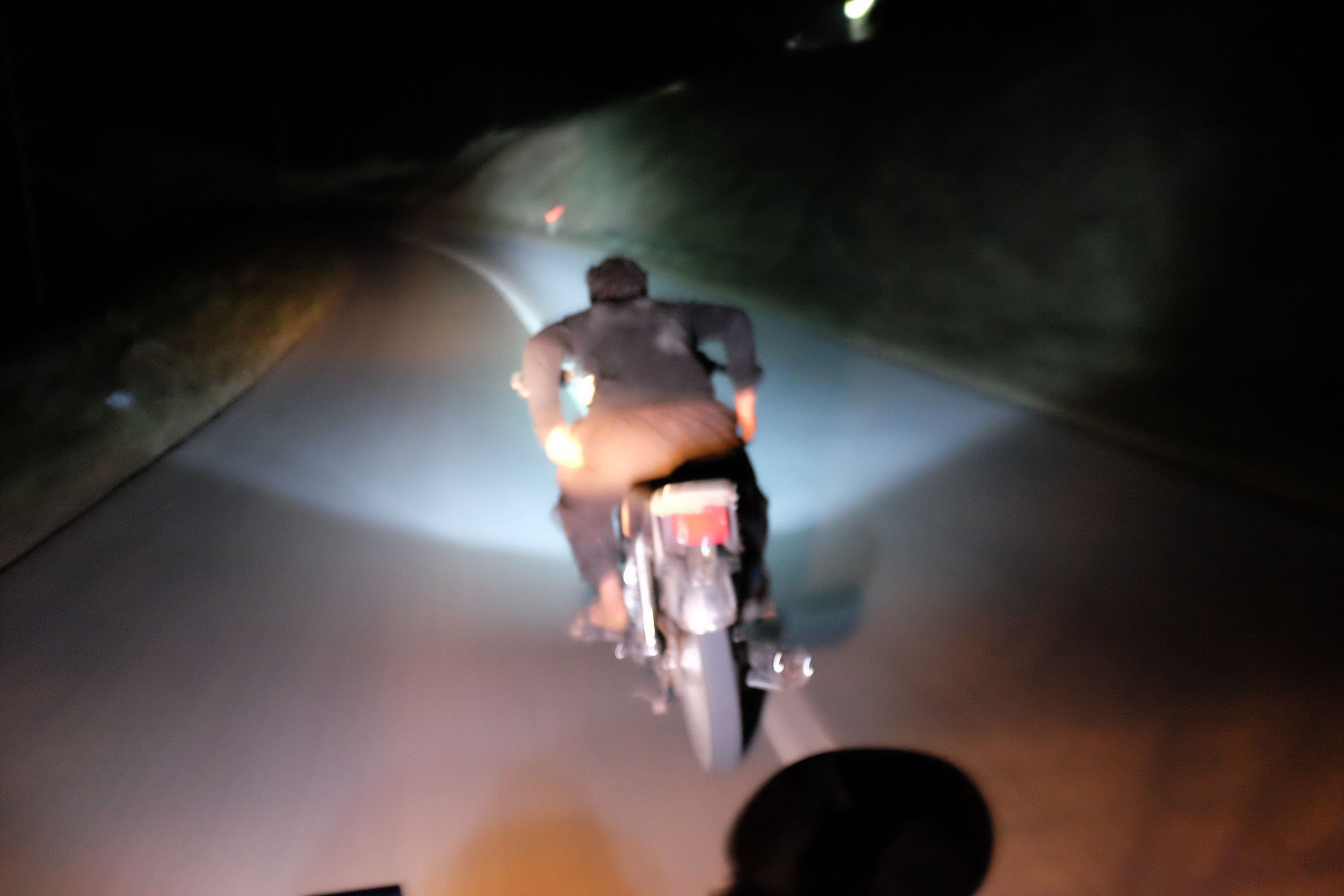A man in traditional Kurdish dress rides a motorcycle at night without holding on to its handlebars, as seen from another motorcycle.