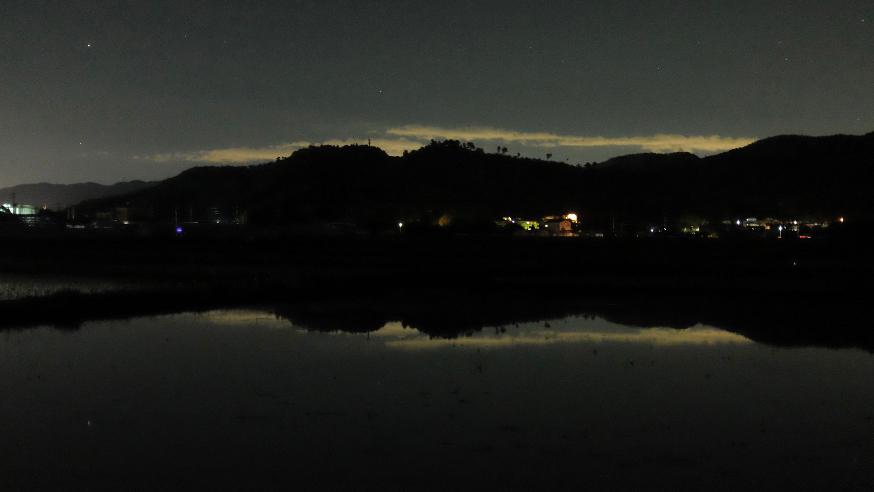 The glow of a city in the night sky is visible over low hills beyond flooded rice paddies.