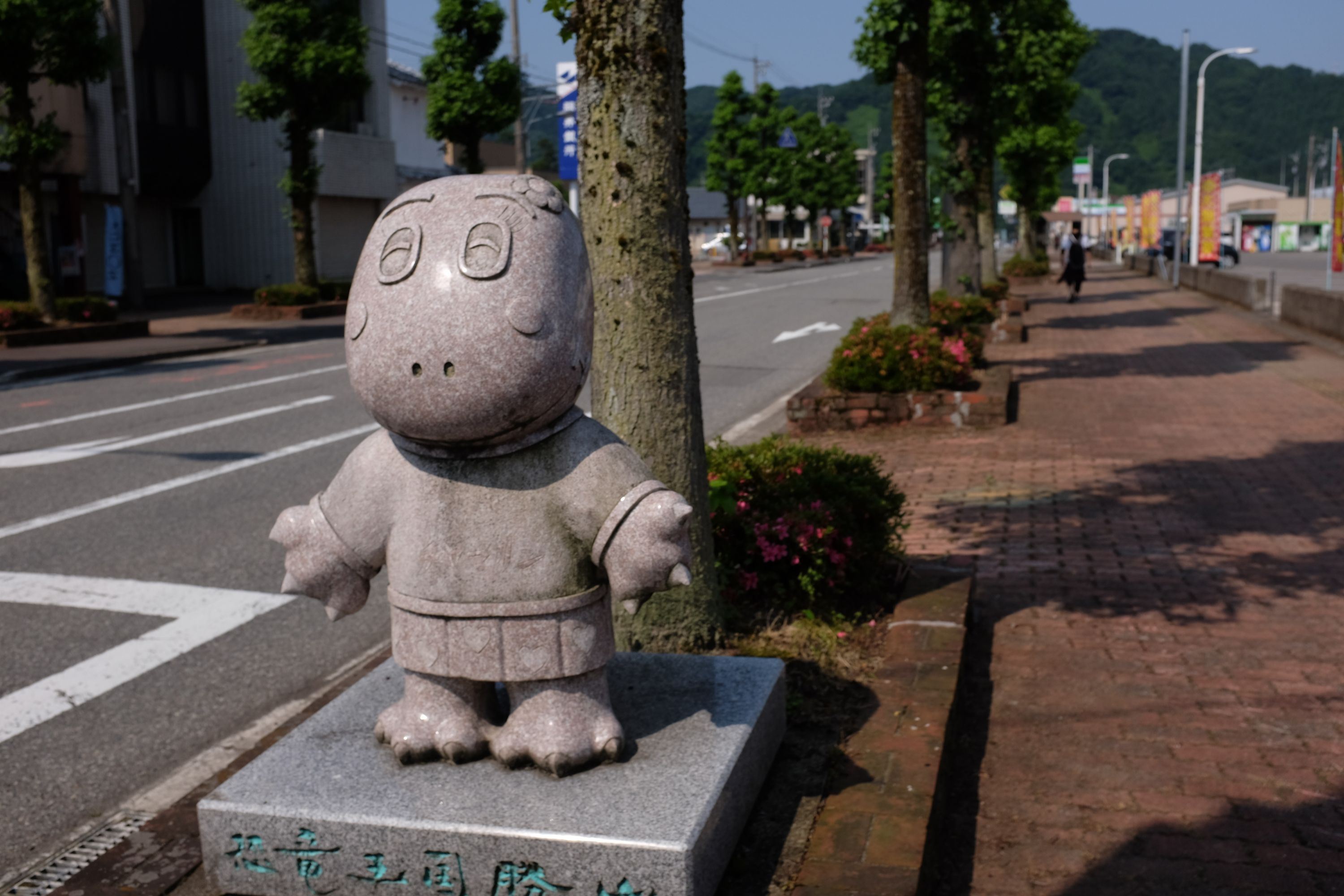 Stone statue of a cartoon-like dinosaur on a sunlit street.