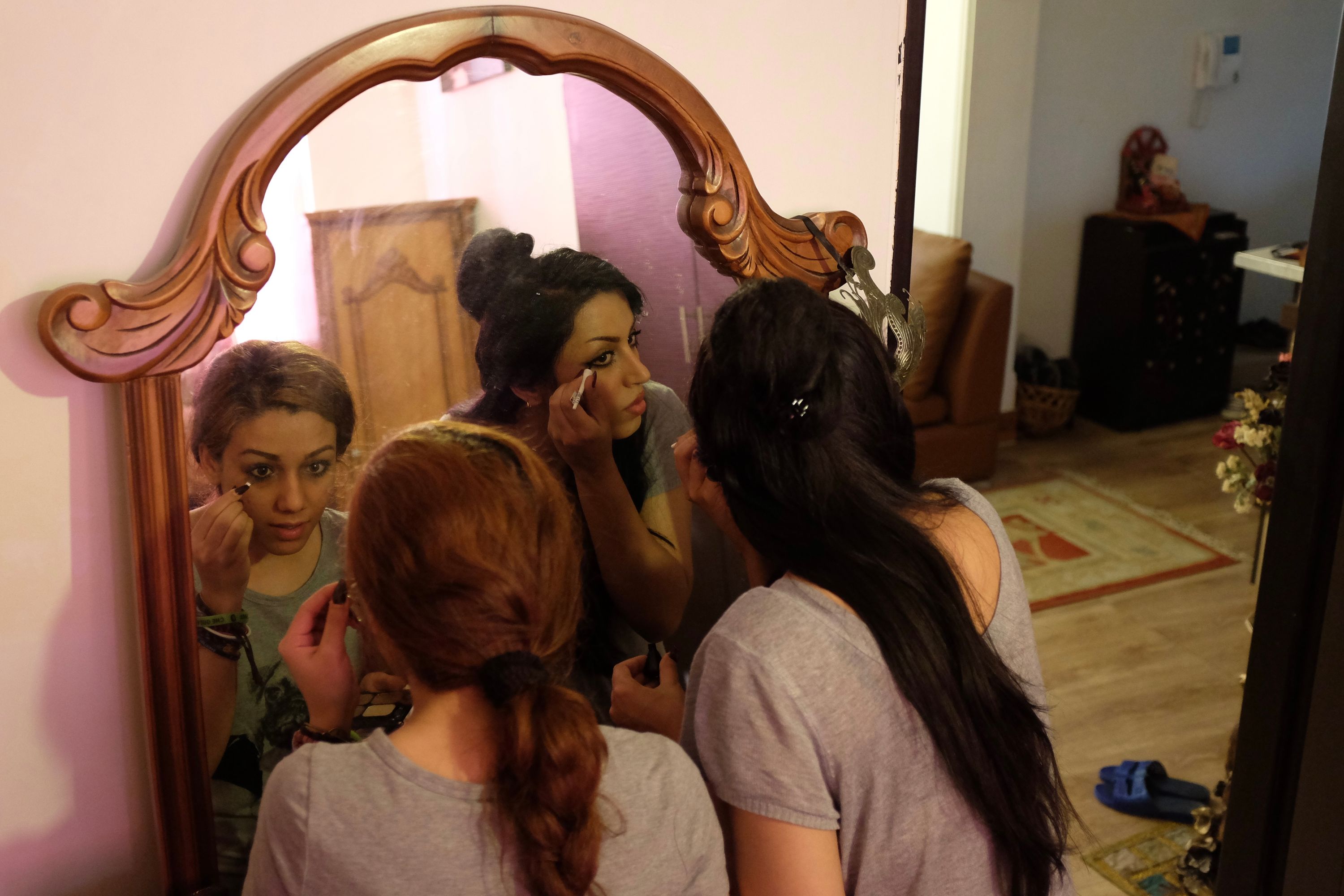 The black-haired woman is joined by a brown-haired woman in front of a mirror, where they apply makeup together.