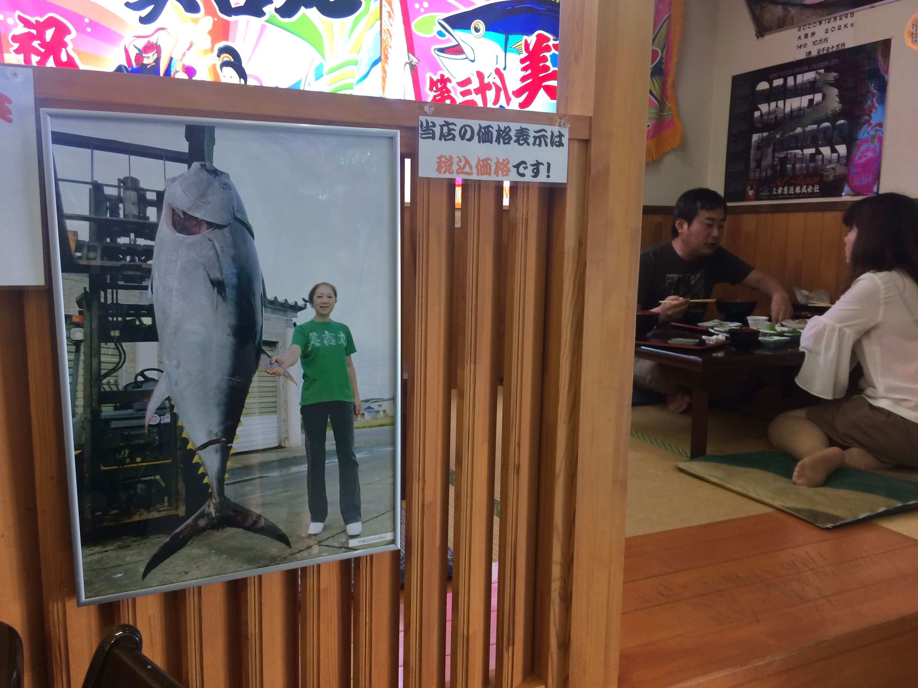 View of a seafood restaurant with two customers at a table, and a picture of a woman posing with a bluefin tuna much larger than she is in the foreground.