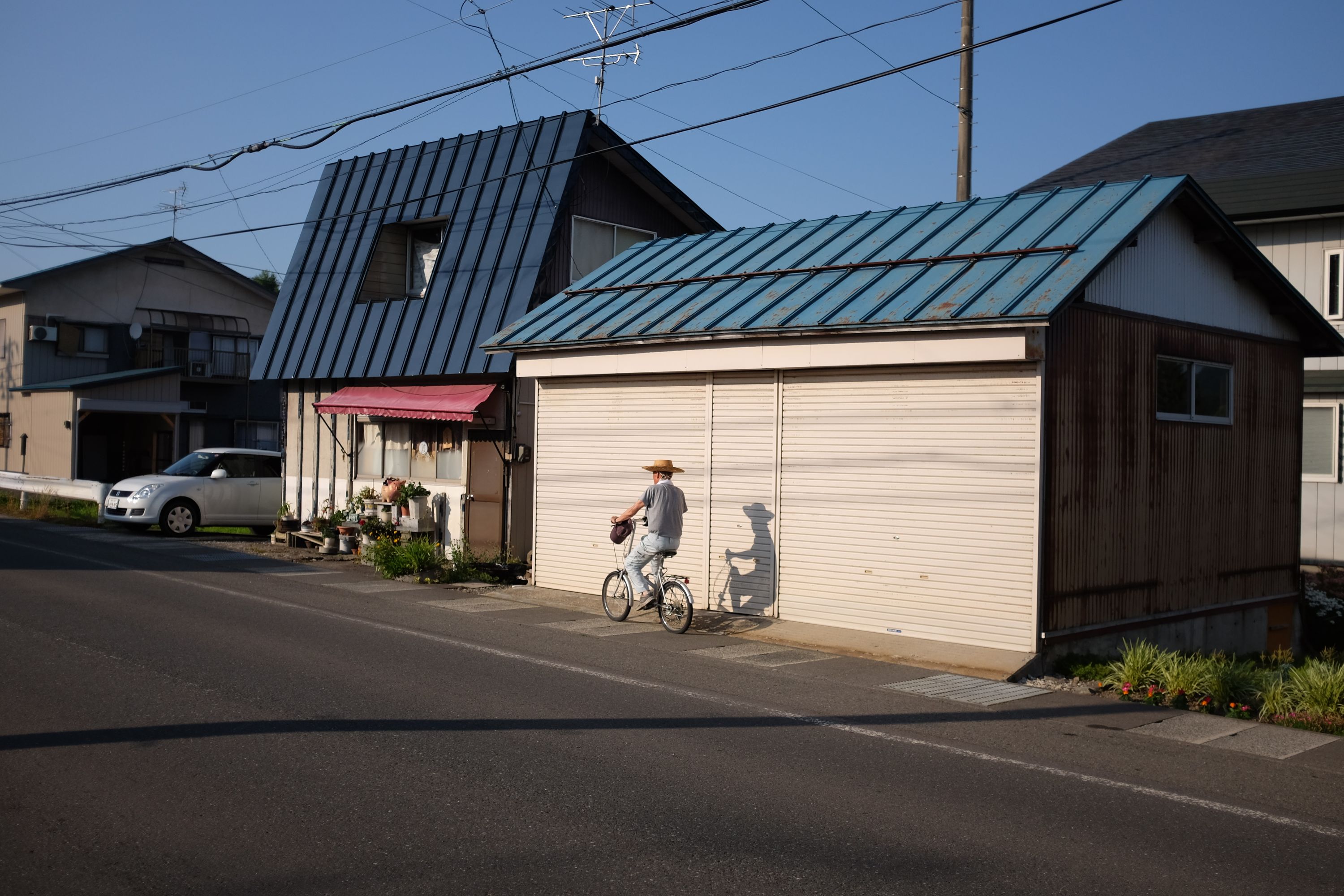 A man in a straw hat rides a bicycle in front of a house with a steep, asymmetrically pitched roof.