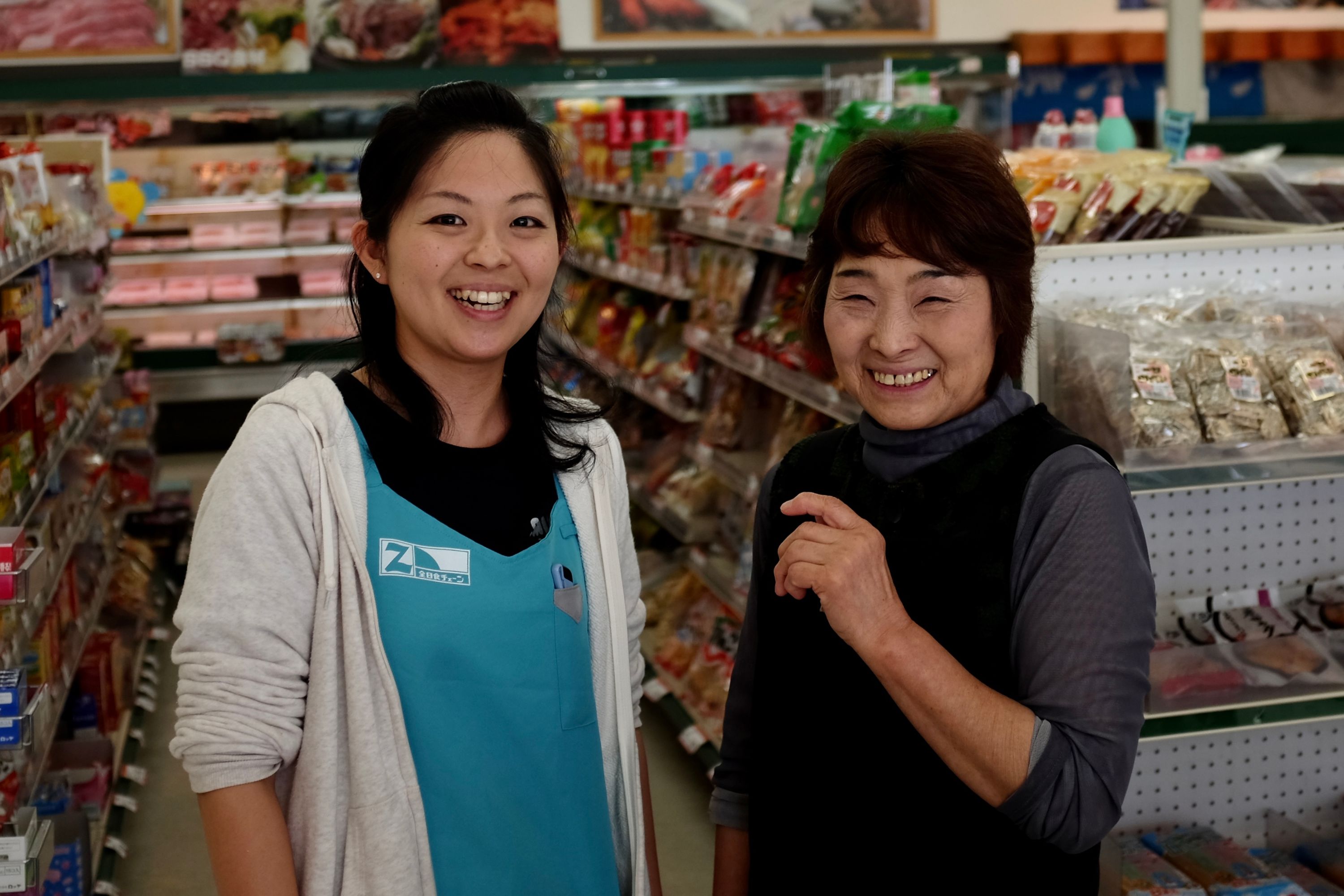 Two shopkeepers, a younger and an older woman, smile into the camera in their shop.