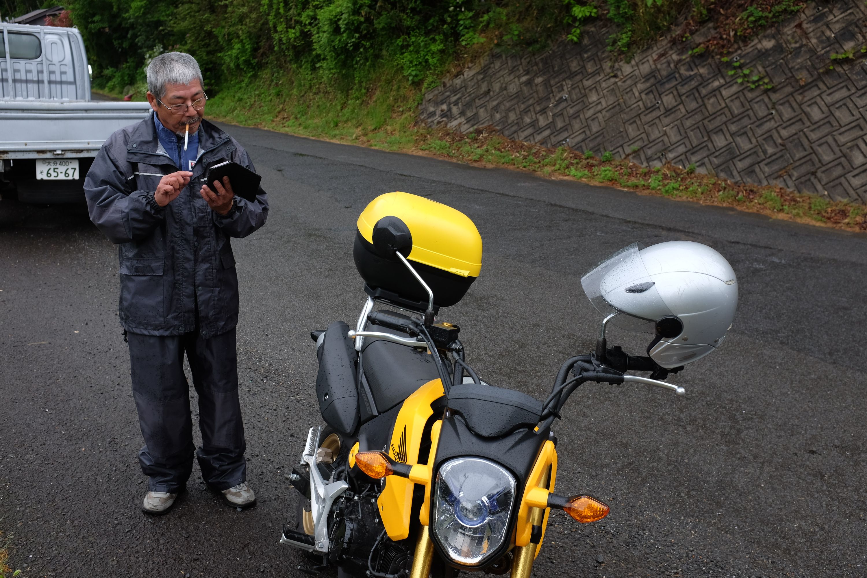 A Japanese man in mechanic’s overalls stands by his yellow motorcycle in the rain, smoking a cigarette and typing into his phone.
