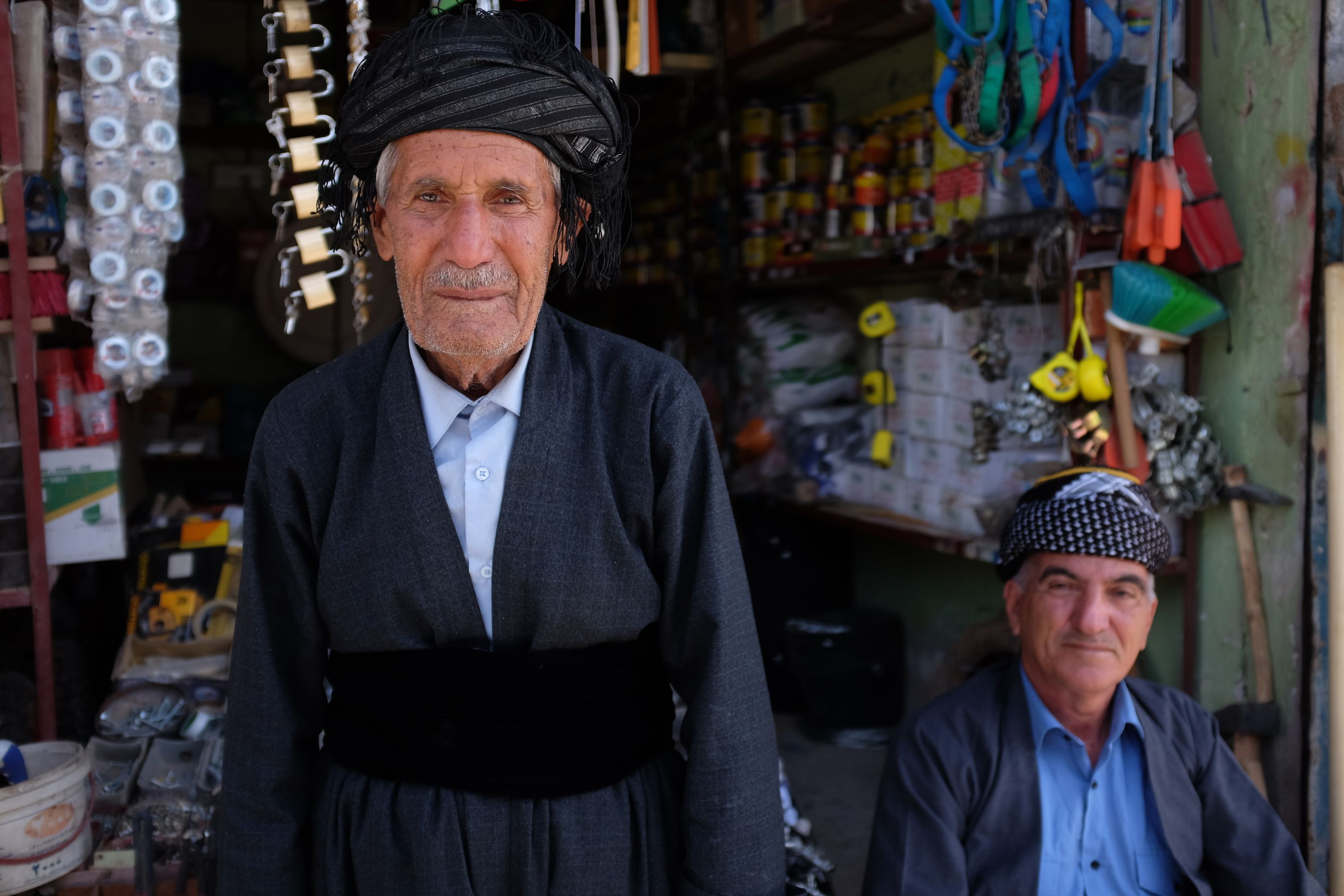 Two men in traditional Kurdish dress pose in front of a general store.