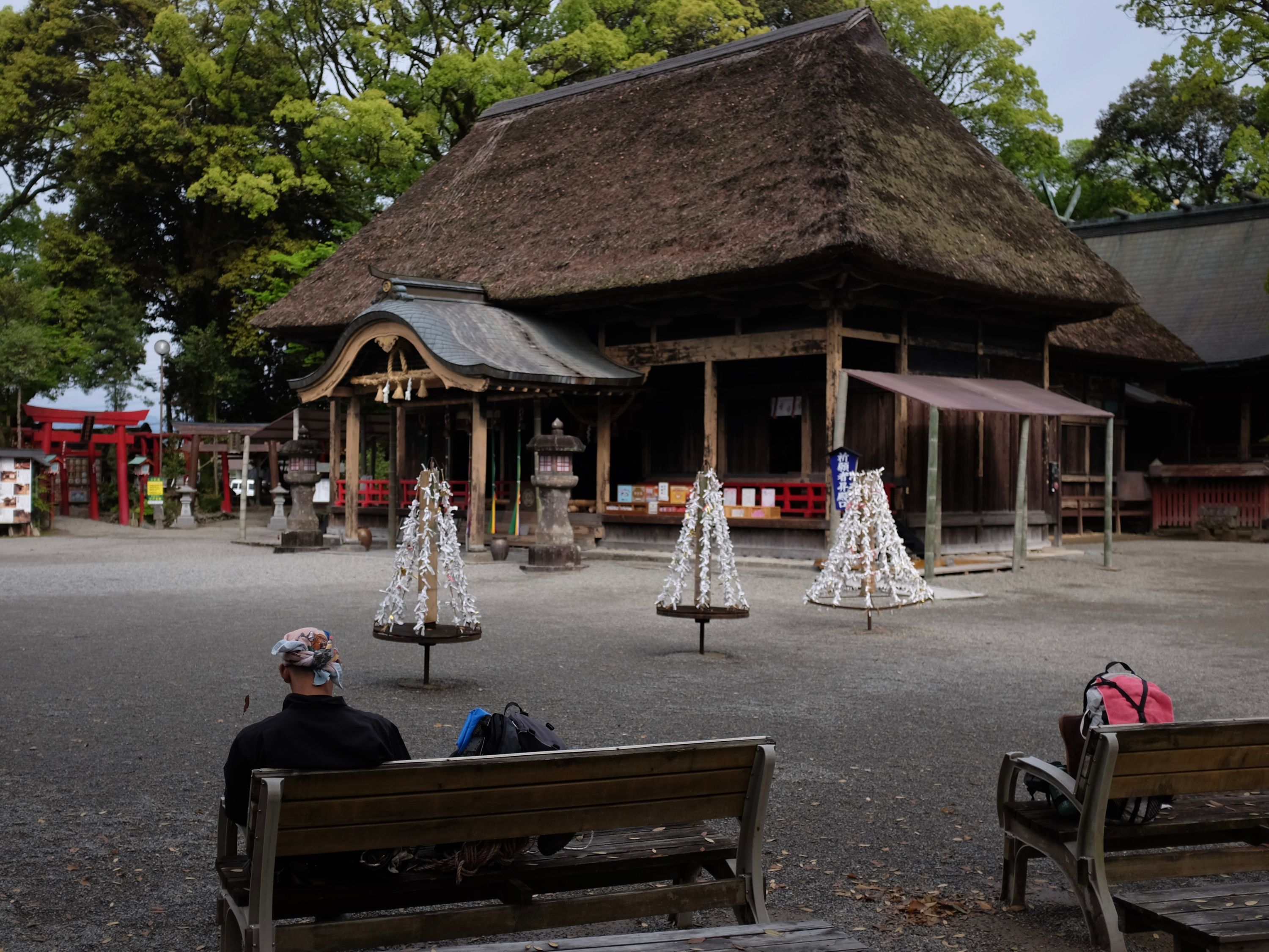 A man sits on a bench in the courtyard of a large, thatch-roofed shrine.